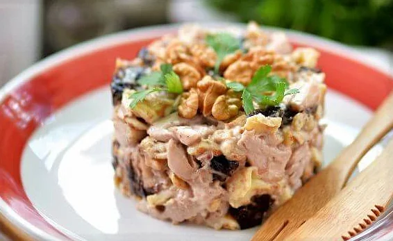 Выложенный с помощью формовочного кольца салат с курицей, черносливом и куриными блинчиками на белой тарелке с красным ободом и деревянными столовыми приборами