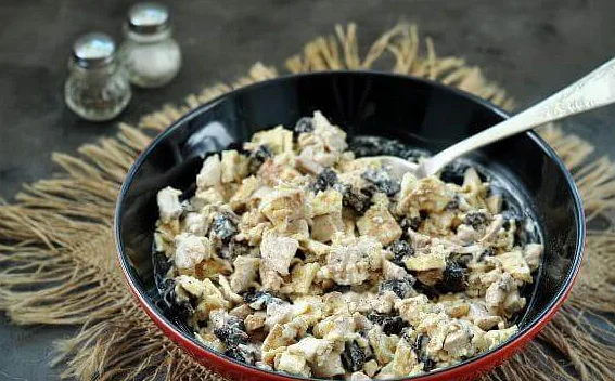 Салат с курицей, черносливом и яичными блинчиками в миске с ложкой на столе